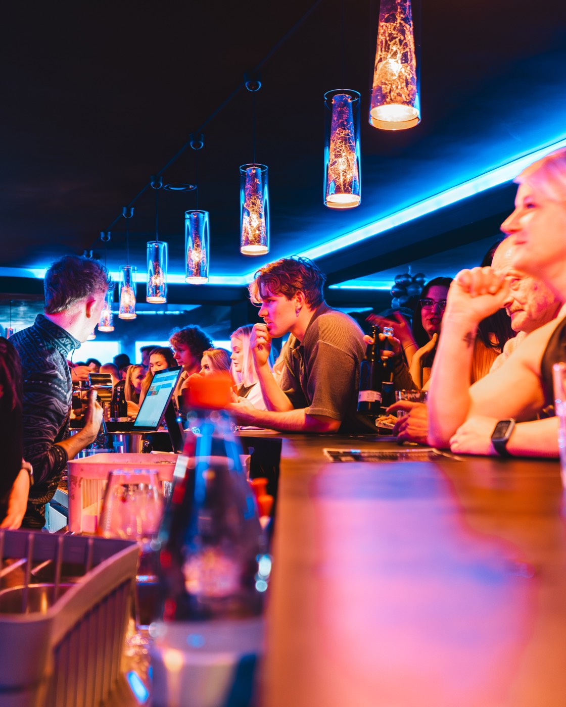 Gäste an der Bar im Nexxus Club, Barkeeper im Einsatz, warme Hängeleuchten vor blauer LED-Leiste
