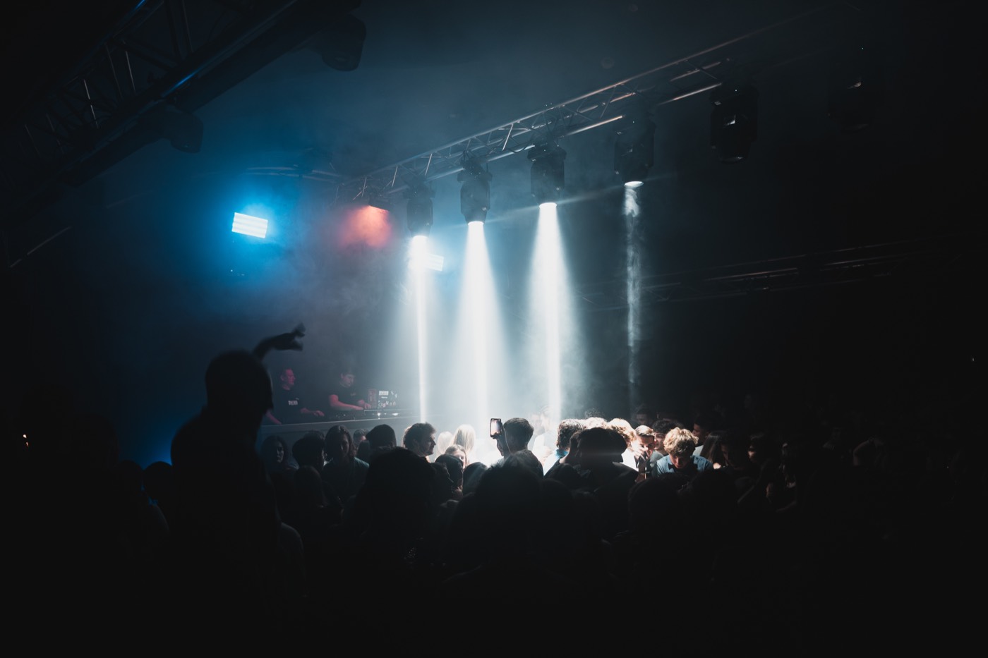 Tanzende Menge im Nebel, weiße Scheinwerferbahnen von der Decke, DJ-Pult im Hintergrund
